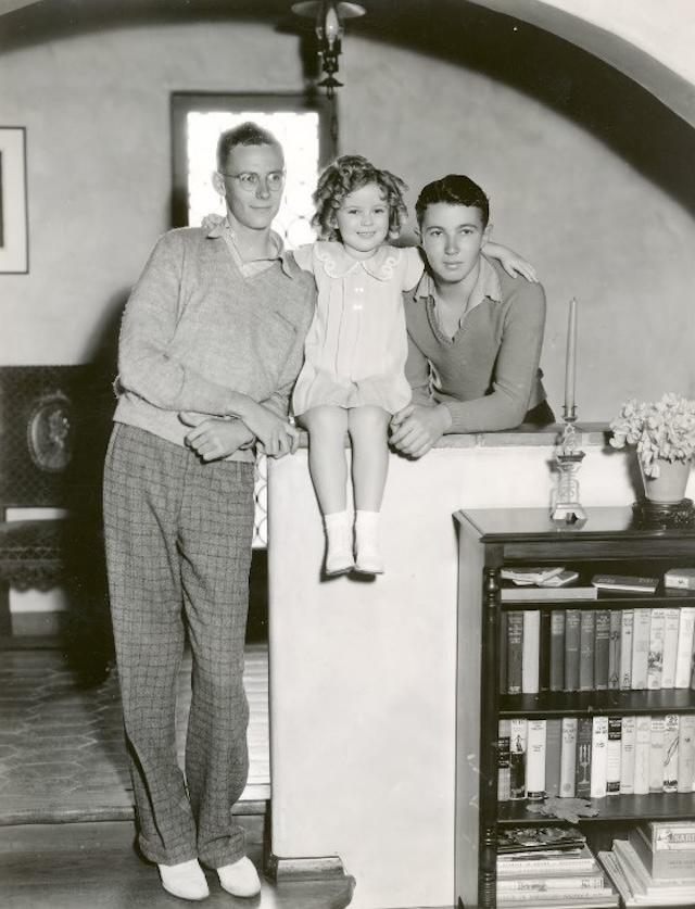 Shirley Temple sits joyfully between her two older brothers, John and George Jr., during a summer day in their backyard, all smiling and soaking up the sun